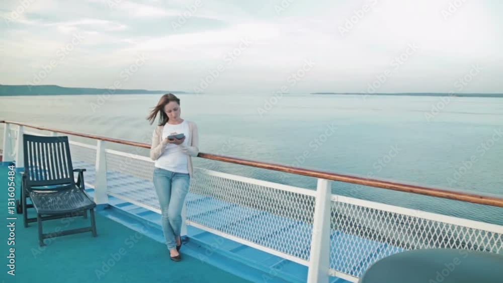 Woman using tablet on deck of cruise ship. Scrolling and touching. Sunrise light, golden hour. Wind waving her hair. The river and riverside can be seen in the background behind her. Woman using tablet on deck of cruise ship. Scrolling and touching. Sunrise light, golden hour. Wind waving her hair. The river and riverside can be seen in the background behind her.