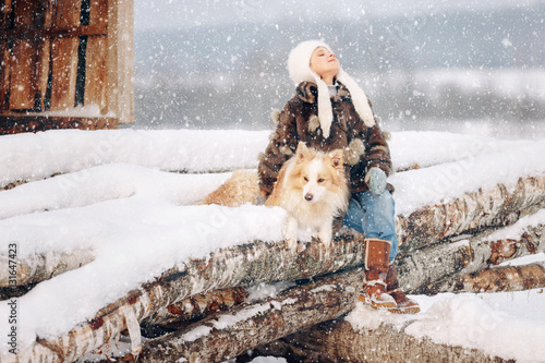 Cute lovely girl child in a fur coat and hat sitting on logs in the winter next to his beloved dog hugs her around and goes soft snow