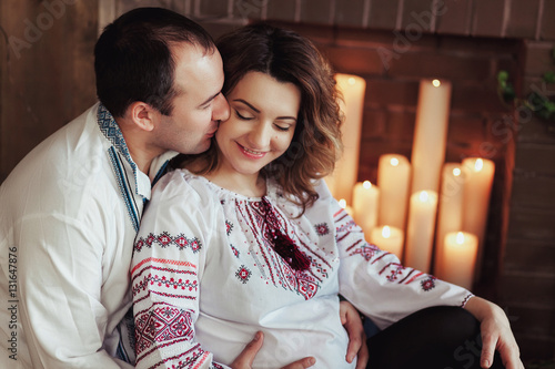 Beautiful young pregnant couple dressed in national ukrainian style sitting by the fireplace with candles. Maternity and family happiness concept photo