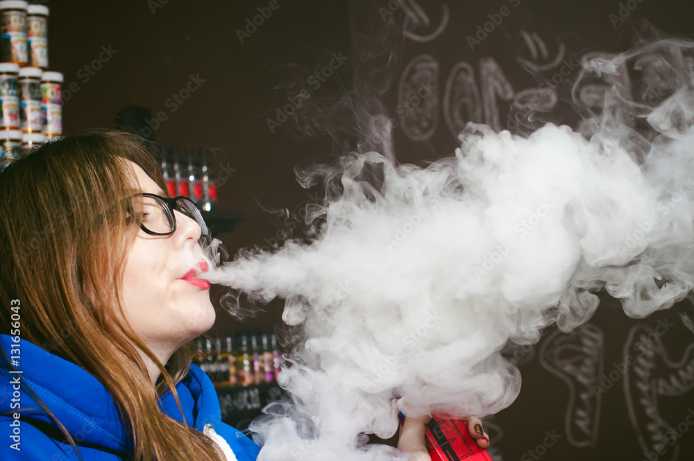 young Cute woman, the seller in vape shop shop, smoking electronic cigarette,  vaping and releases a cloud of vapor