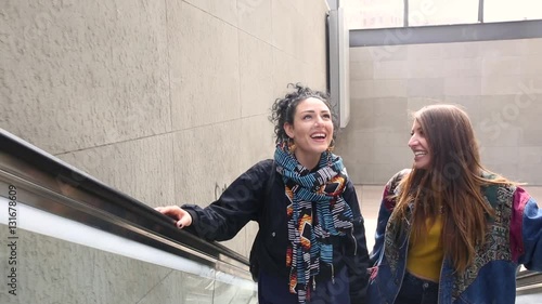 Lesbian couple using an escalator in Potsdamer Platz, Berlin. Two beautiful women on their twenties walking and embracing. Close up shot, head and shoulders view. Homosexuality and lifestyle concepts.