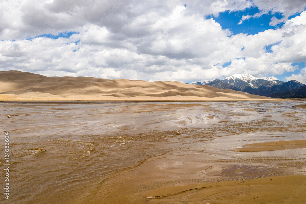 Spring view of gently flowing Medano Creek at base of rolling sand dunes and snow-capped Mt Herard. Great Sand Dunes National Park & Preserve, Colorado, USA.