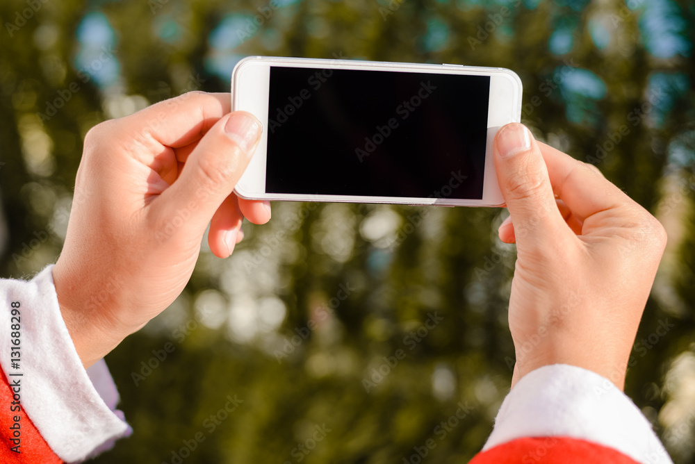 Santa Claus holding using mobile smartphone ready for Christmas time on green fir background Santa Claus holding using mobile smartphone ready for Christmas time on green fir background
