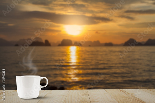 Hot coffee cup with steam on wooden table top on blurred golden sky, sea and island background during sunrise © shark749