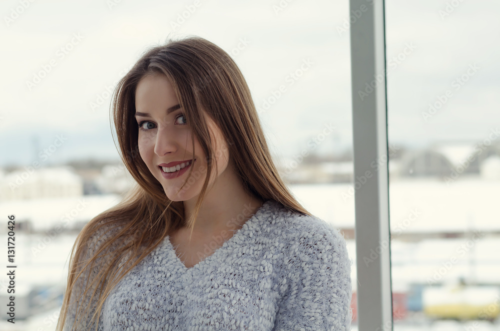 Photo of young beautiful happy smiling woman with long hair near the window. Girl looking at the camera. Female fashion. Sunny day. City lifestyle. Waist up. Toned