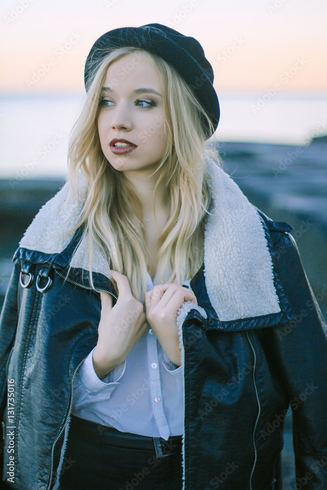 Obraz premium beautiful young blond woman standing by the sea at sunset wearing a black hat, a sheepskin coat and a white shirt. She is looking into the distance