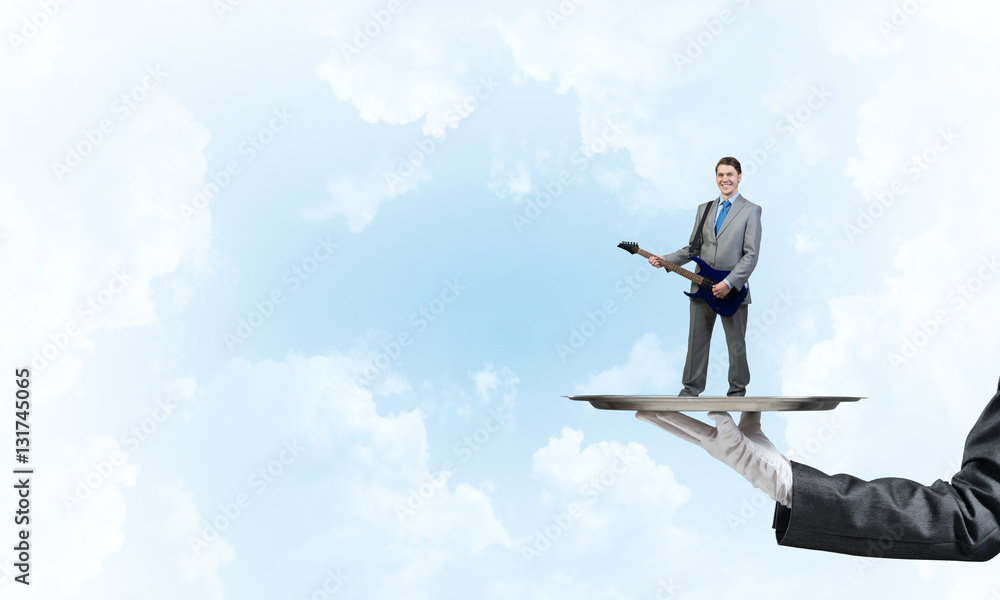 Businessman on metal tray playing electric guitar against blue sky background Businessman on metal tray playing electric guitar against blue sky background