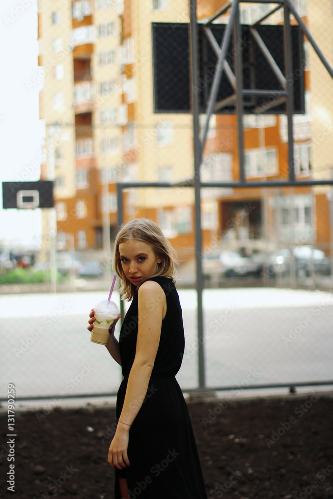 girl in a black dress with a cocktail in a residential area on the background of basketball field  