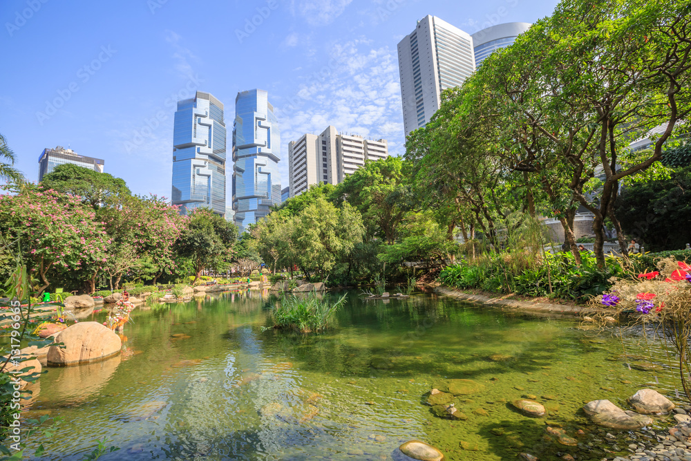 Fototapeta premium Beautiful view of pond at the lush Hong Kong Park surrounded by skyscrapers in the Central business district in Hong Kong island. Sunny day with blue sky.