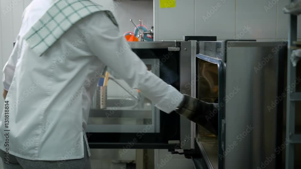 Chef wearing a white uniform opens the oven door pulls out a tray of freshly baked rolls. Preparation of delicious pastries on a restaurant kitchen.