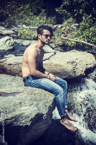 Athletic shirtless young man outdoor at river or water stream, sitting on big rock, looking away, with stones in background