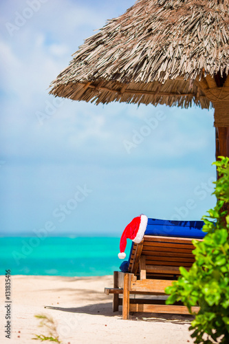 Santa Claus Hat on chair near tropical beach with turquoise sea water and white sand. Christmas vacation concept