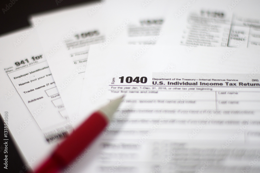 Tax Season: Horizontal left side view of 1040 U.S. Individual Income Tax Return Form with an office laptop background with red white pen and eye glasses foreground Tax Season: Horizontal left side view of 1040 U.S. Individual Income Tax Return Form with an office laptop background with red white pen and eye glasses foreground