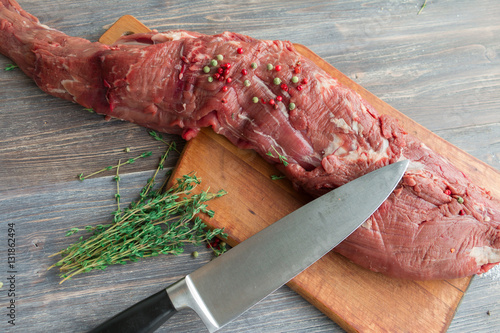 Raw red beef tender loin on wooden kitchen board with green and pink pepper and thyme. Selective focus. Top view.