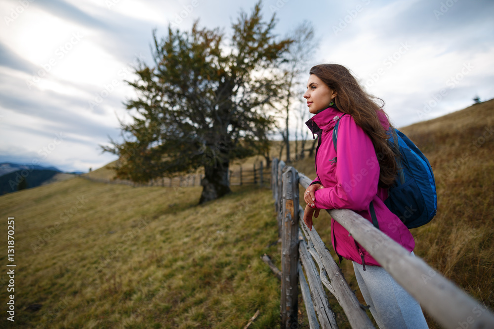 A hiker stands near a wooden fence and enjoy mountain valley. Shot of a young woman looking at the landscape while hiking in the mountains