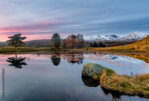 Calm pink sunset with reflections at Kelly Hall Tarn in the Lake District with snow on the Old Man Of Coniston. photo