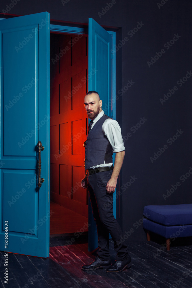 Stylish young fashion model in a suit near door with red light. Business style. Fashionable image. Evening dress. Sexy man standing and looking at the camera. Fashion look.
