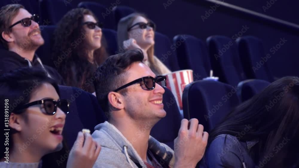 Young people in 3D viewers laughing at the movie theater. Caucasian brunette girl touching her hair at the cinema. Asian girl taking flake of popcorn from the bucket of caucasian guy Young people in 3D viewers laughing at the movie theater. Caucasian brunette girl touching her hair at the cinema. Asian girl taking flake of popcorn from the bucket of caucasian guy