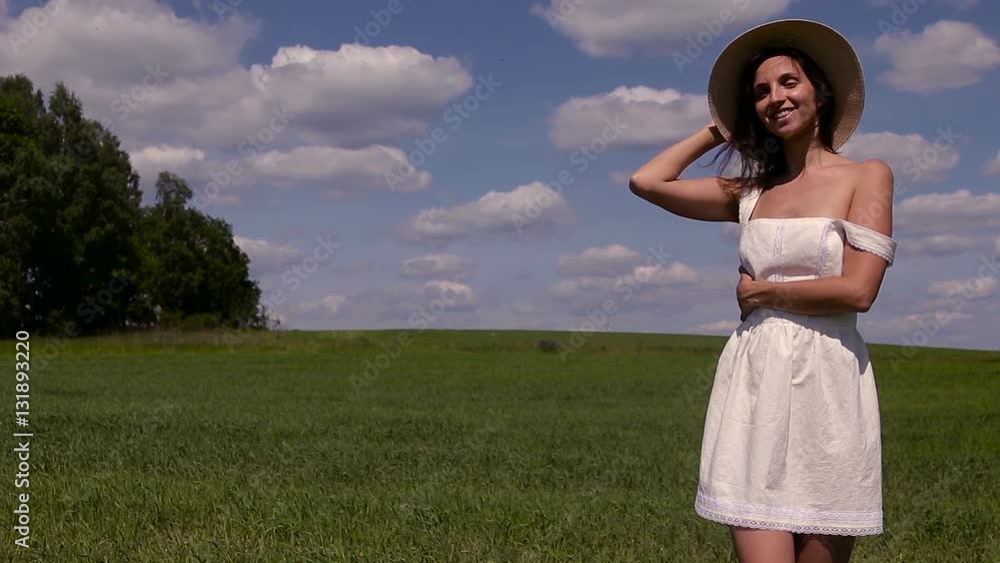 young girl with long black hair stands on the site in white dress and clothes straw hat is on the green field with grass and smiles, summer. young girl with long black hair stands on the site in white dress and clothes straw hat is on the green field with grass and smiles, summer.