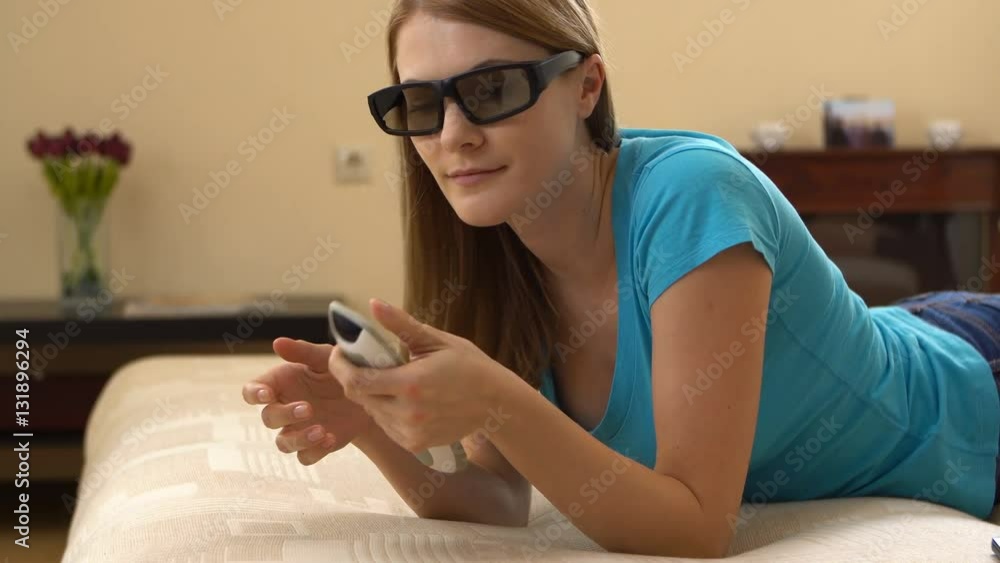 Beautiful attractive young woman in blue t-shirt lying on sofa and watching TV in 3D-glasses. Beautiful attractive young woman in blue t-shirt lying on sofa and watching TV in 3D-glasses.