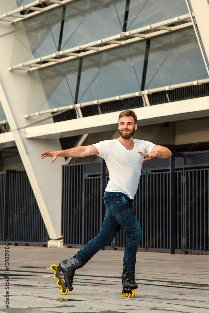 Bearded man on rollerblades standing in building background. Young fit man with white t-shirts and jeans on roller skates riding outdoors after rain.
