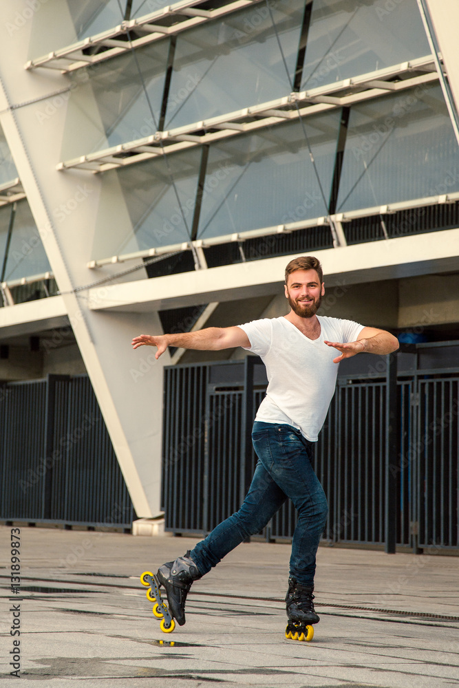 Bearded man on rollerblades standing in building background. Young fit man with white t-shirts and jeans on roller skates riding outdoors after rain.
