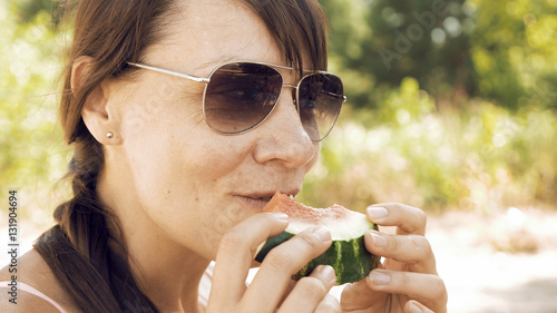 Smiley woman eats an watermelon outdoors on summer sunny day. Eating fruits outdoors on a picnic is a popular way of spending weekends. The female is wearing sunglasses to protect her from sun.