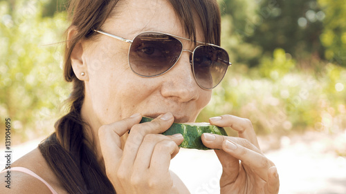 Smiley woman eats an watermelon outdoors on summer sunny day. Eating fruits outdoors on a picnic is a popular way of spending weekends. The female is wearing sunglasses to protect her from sun.