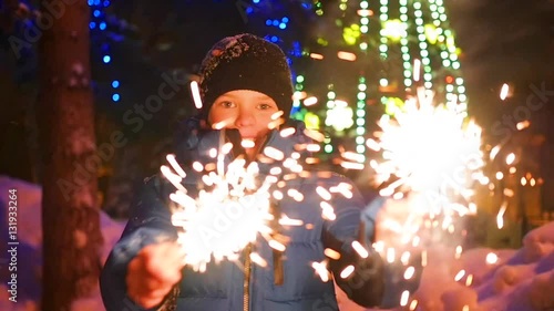 The child holds the sparklers outdoors in the winter. Slowmotion . In the background, lights and garlands of Christmas fir