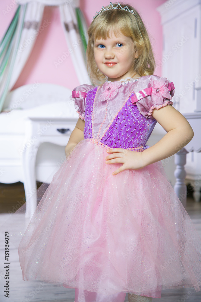 Little happy princess girl in pink dress and crown in her royal room posing and smiling.
Little happy princess girl in pink dress and crown in her royal room posing and smiling.