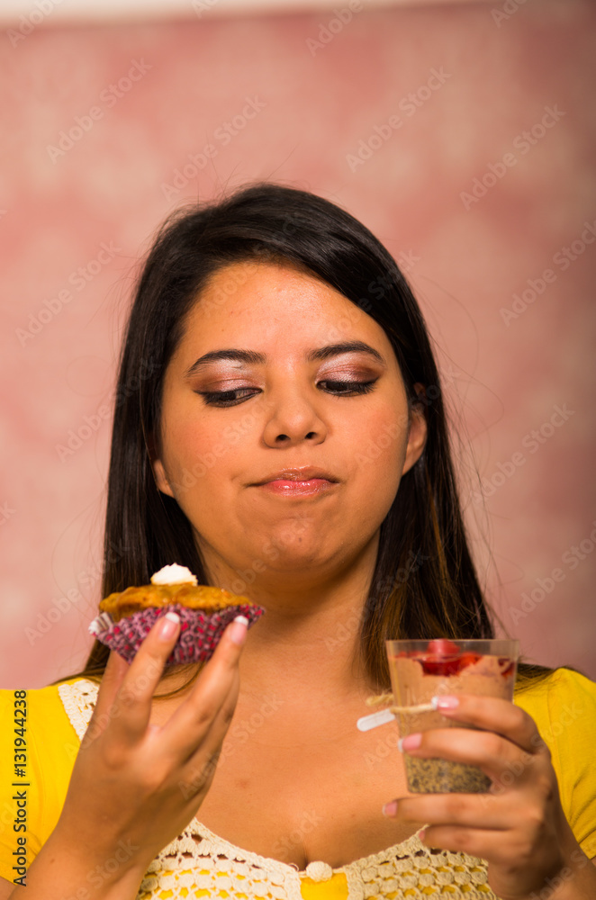 Brunette woman holding delicious brown colored muffin with cream topping, glass of mousse in other hand, big smile and ready to take a bite, pastry concept Brunette woman holding delicious brown colored muffin with cream topping, glass of mousse in other hand, big smile and ready to take a bite, pastry concept