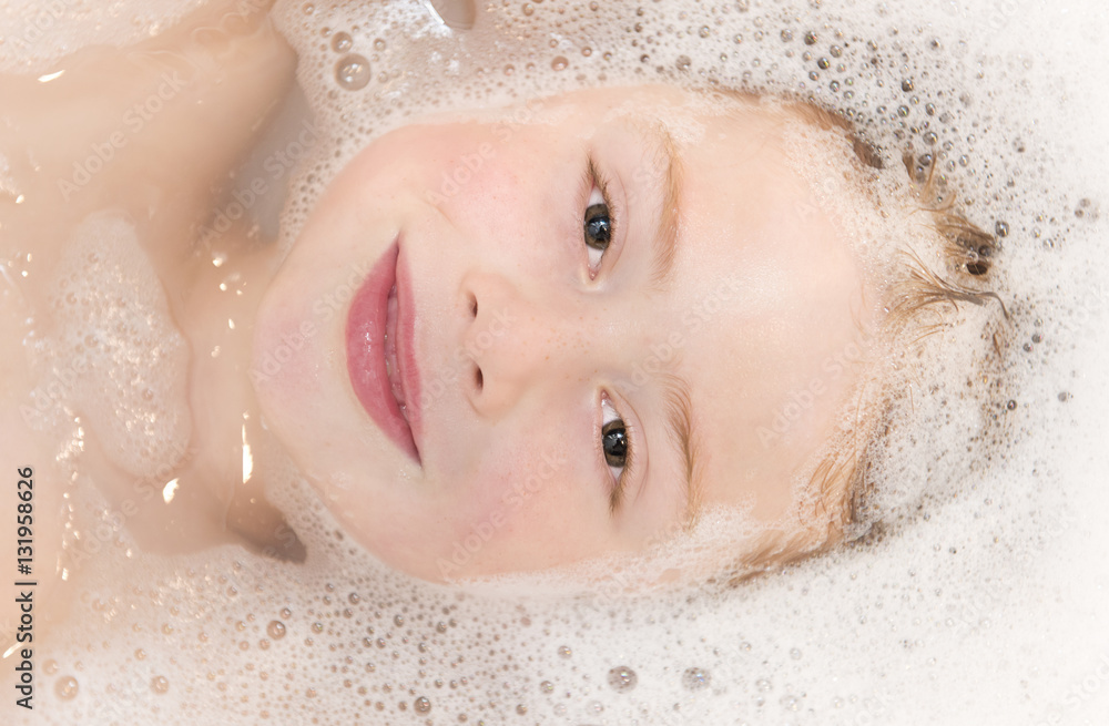 Fototapeta premium Cute ginger haired boy lying in the bath which is fullof bubbles. His head is prtly submerged and he is looking into the camera. Upper body shot includes head and shoulders.