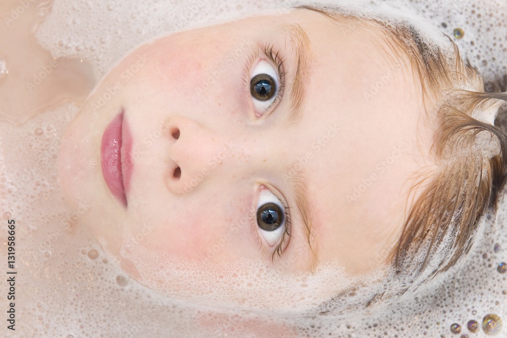 Naklejka premium Close up shot of cute ginger haired boy lying in the bath. He is looking directly into the camera and his head is partly submerged in the water which has bubbles in it. 