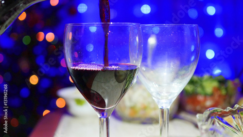 Red wine is poured into glasses. In the background, bokeh lights and garlands of Christmas fir