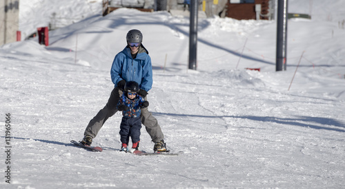 Toddler Learning to Ski with his Father at a Colorado Resort. Dressed Safely with Helmet, Sunglasses, & Harness.