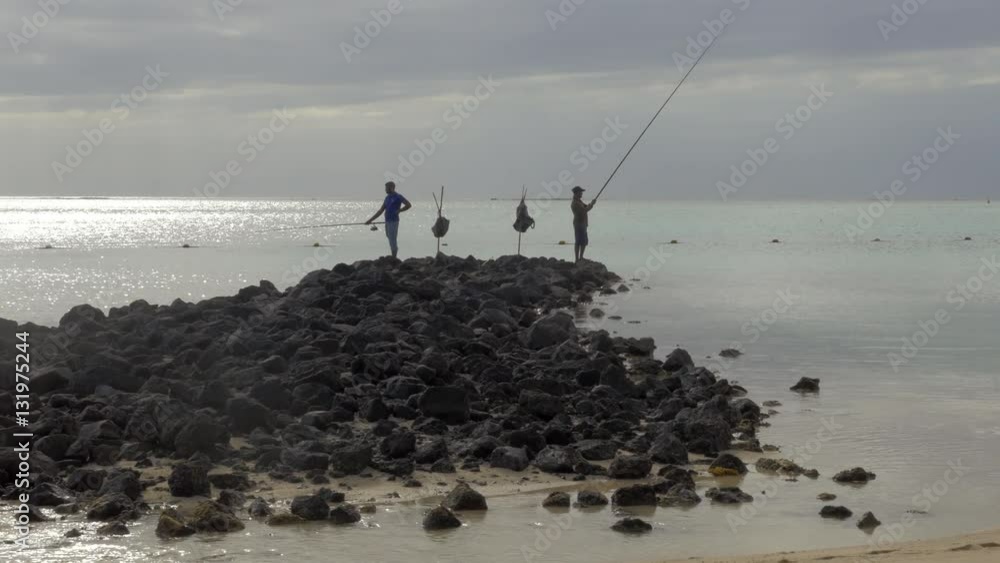MAURITIUS - JUNE 17, 2016: Two men standing on the rocks on the coast and fishing. Scene with cloudy sky and quiet ocean MAURITIUS - JUNE 17, 2016: Two men standing on the rocks on the coast and fishing. Scene with cloudy sky and quiet ocean