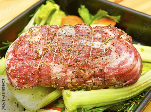 Rolled up raw lamb joint seasoned with salt, pepper and rosemary. Joint is resting on a trivet of celery, onions, carrot and rosemary