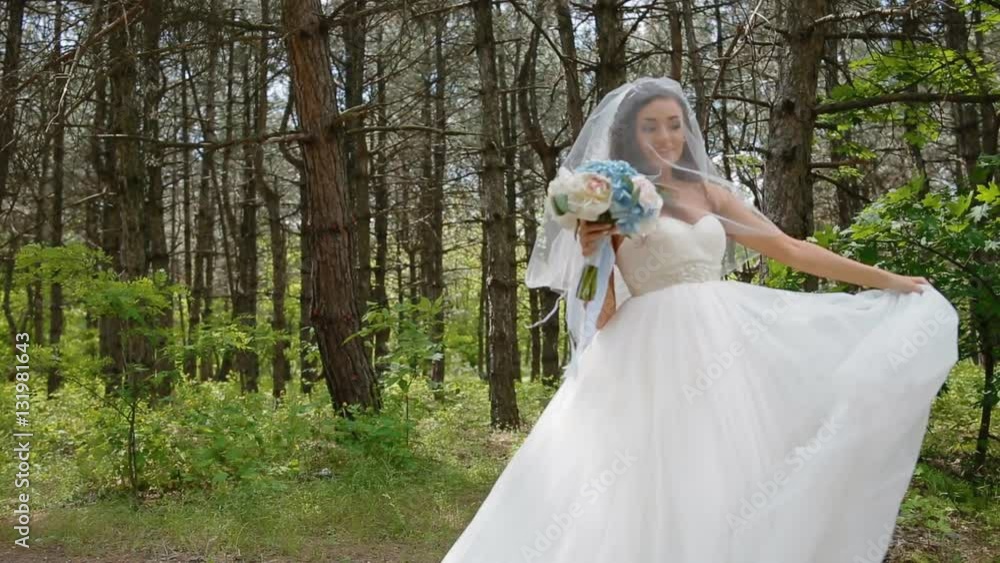 Beautiful bride pose with wedding bouquet outdoor in sunny day. Caucasian young woman with long dark curly hair gently keep the flowers and spinning arounf playing with her fluffy dress. Beautiful bride pose with wedding bouquet outdoor in sunny day. Caucasian young woman with long dark curly hair gently keep the flowers and spinning arounf playing with her fluffy dress.