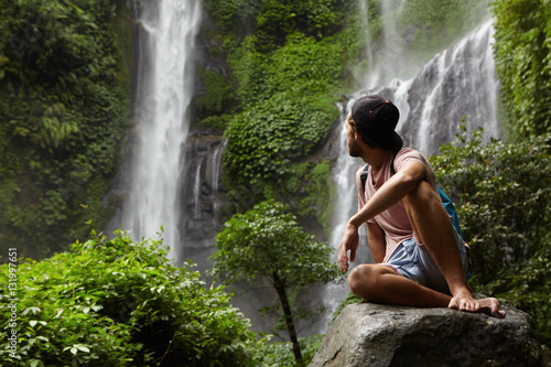 Travel and adventure. Fashionable young man wearing snapback and backpack sitting on stone and looking back at waterfall in beautiful green rainforest. Barefooted tourist having rest on rock in jungle