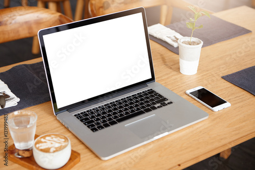 Business, technology and communication concept. Minimalistic workspace with modern laptop computer with white blank screen, generic mobile phone, coffee and glass of water on wooden desk. Mock up