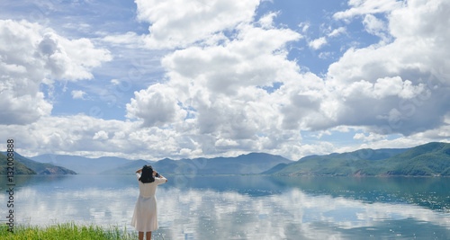 Single young Chinese woman in white long skirt stands on wasted platform on bank of Lugu lake, listening to music, back to camera, at Daluoshui village, Lijiang, China photo