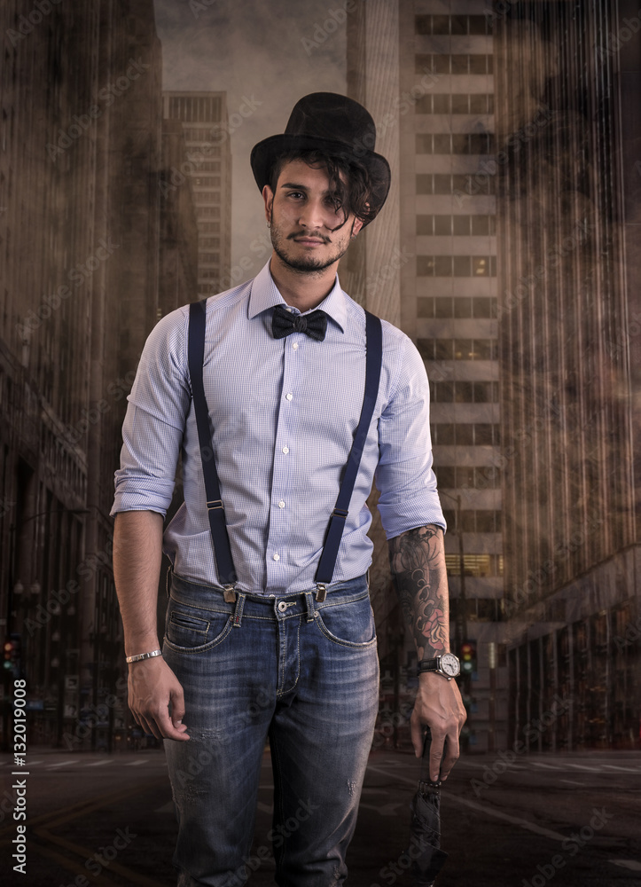 Φωτογραφία Portrait of brunette young man in glasses, hat, bow-tie, suspenders and shirt on dark smoky city street