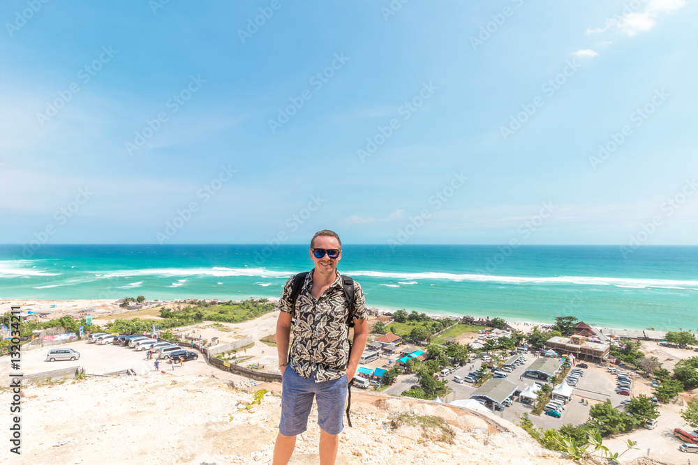 Handsome traveler man stay by blue ocean background - Happy guy relaxing at sea view point - Concept of freedom and summer trip around the world backpacker style. Bali, Indonesia.