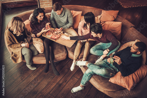 Group of multi ethnic young friends eating pizza in home interior