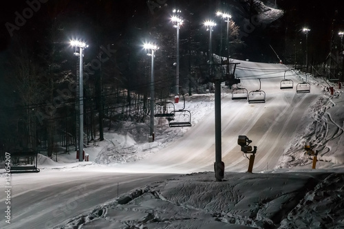 Scenic night view of an illuminated snowy ski track with a chair ski lift. Night skiing service at Sochi Gorky Gorod winter mountain resort