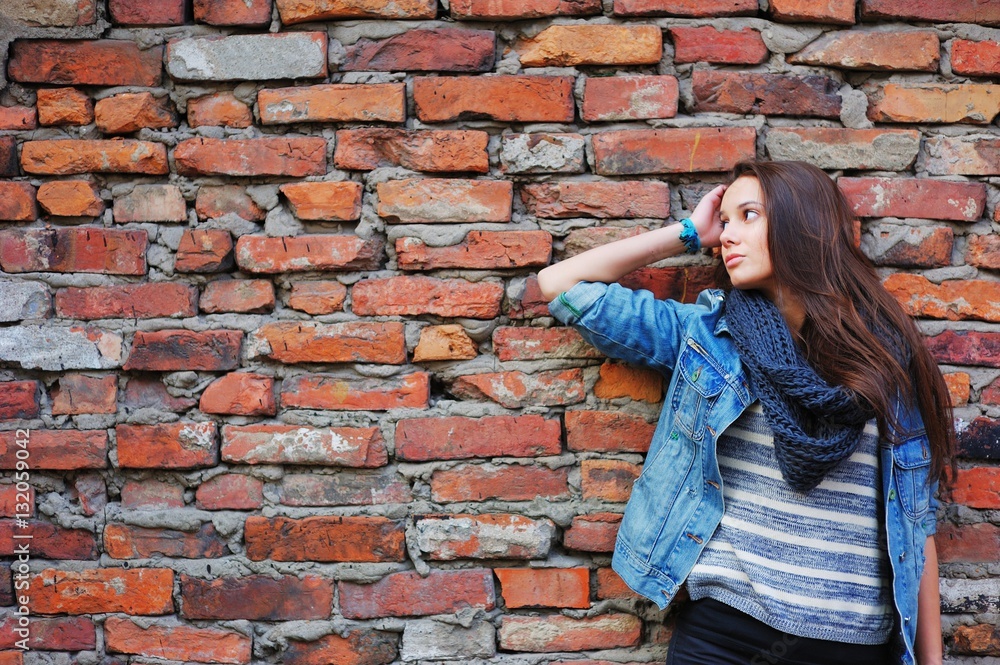 girl with long hair, dressed in a denim jacket and a scarf standing near a wall made of red bricks and waits girl with long hair, dressed in a denim jacket and a scarf standing near a wall made of red bricks and waits