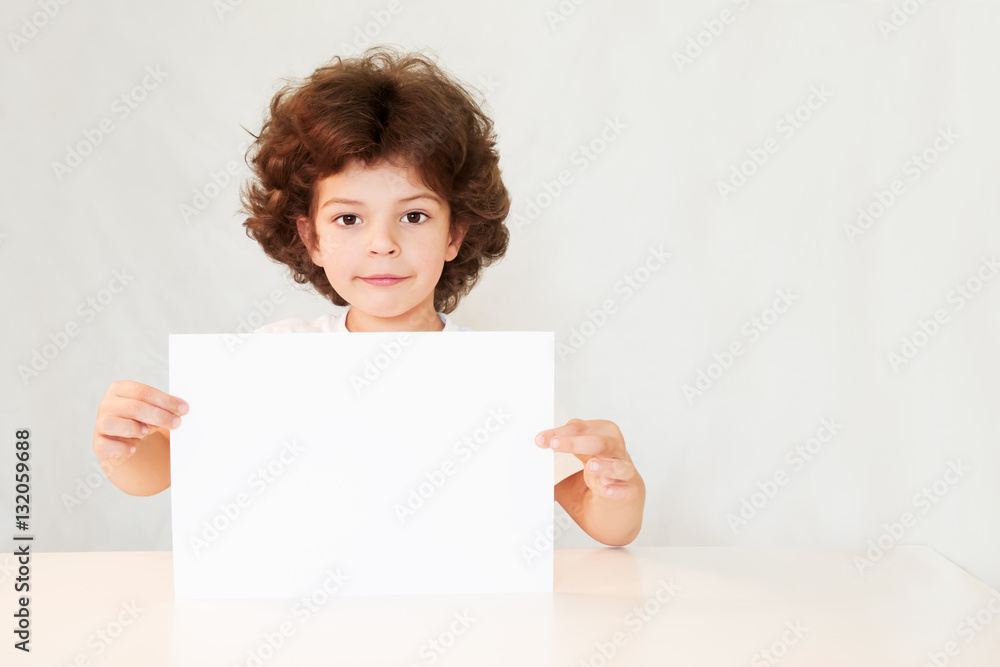 Happy cute little boy holding a blank white sheet of paper with interest and looking at the camera. Gray background. Happy cute little boy holding a blank white sheet of paper with interest and looking at the camera. Gray background.