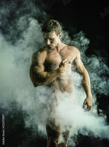 Totally naked male bodybuilder with smoke hiding genitalia, looking away to a side, on dark background