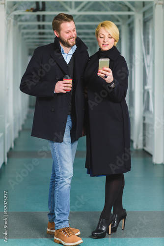 Elegant couple dressed in coat looking on smart phone at lobby
