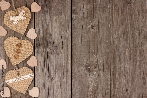 Side border of handmade burlap hearts with ribbon and buttons over a rustic wooden background